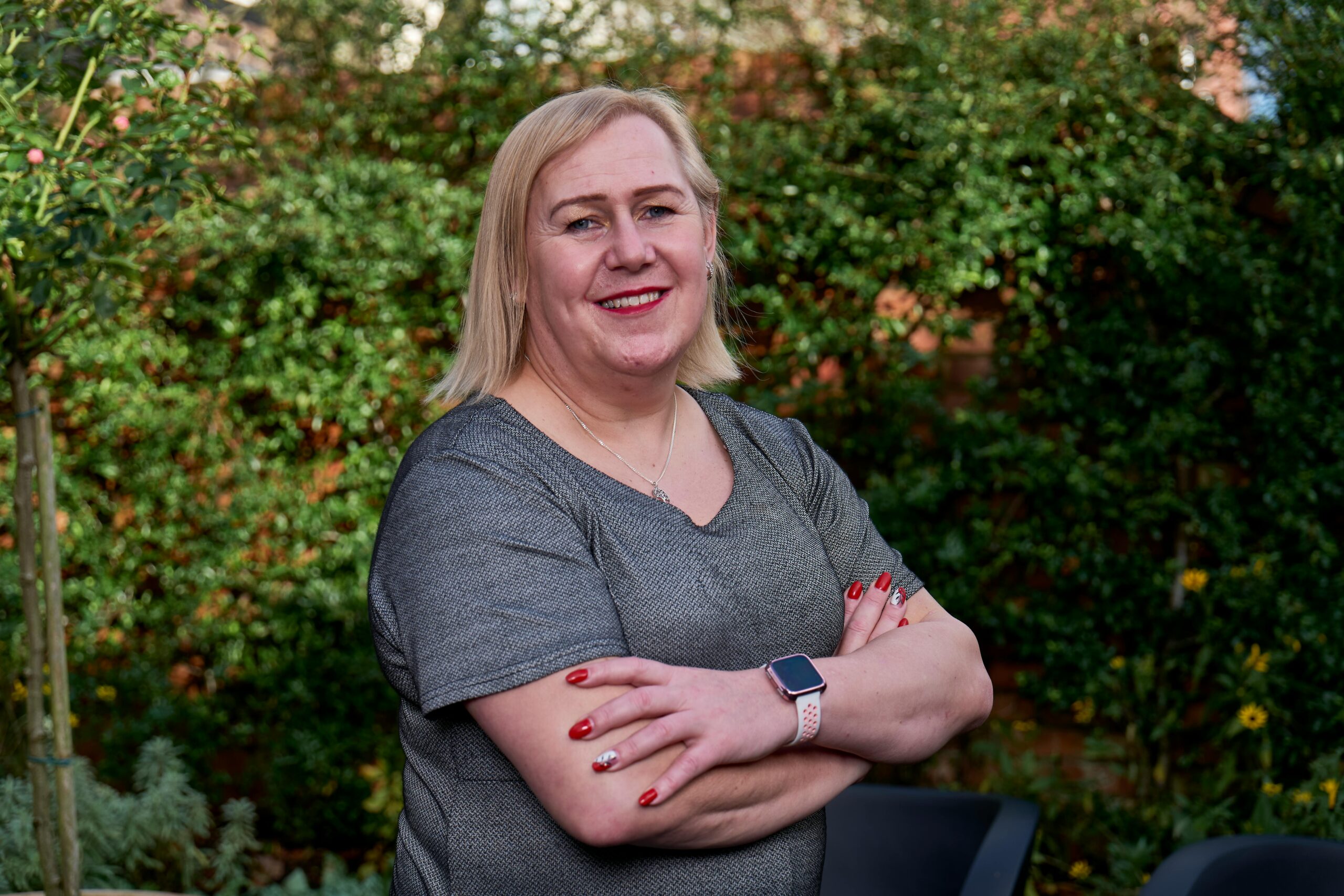 A smiling blonde woman standing outdoors with arms crossed, wearing a smart watch.